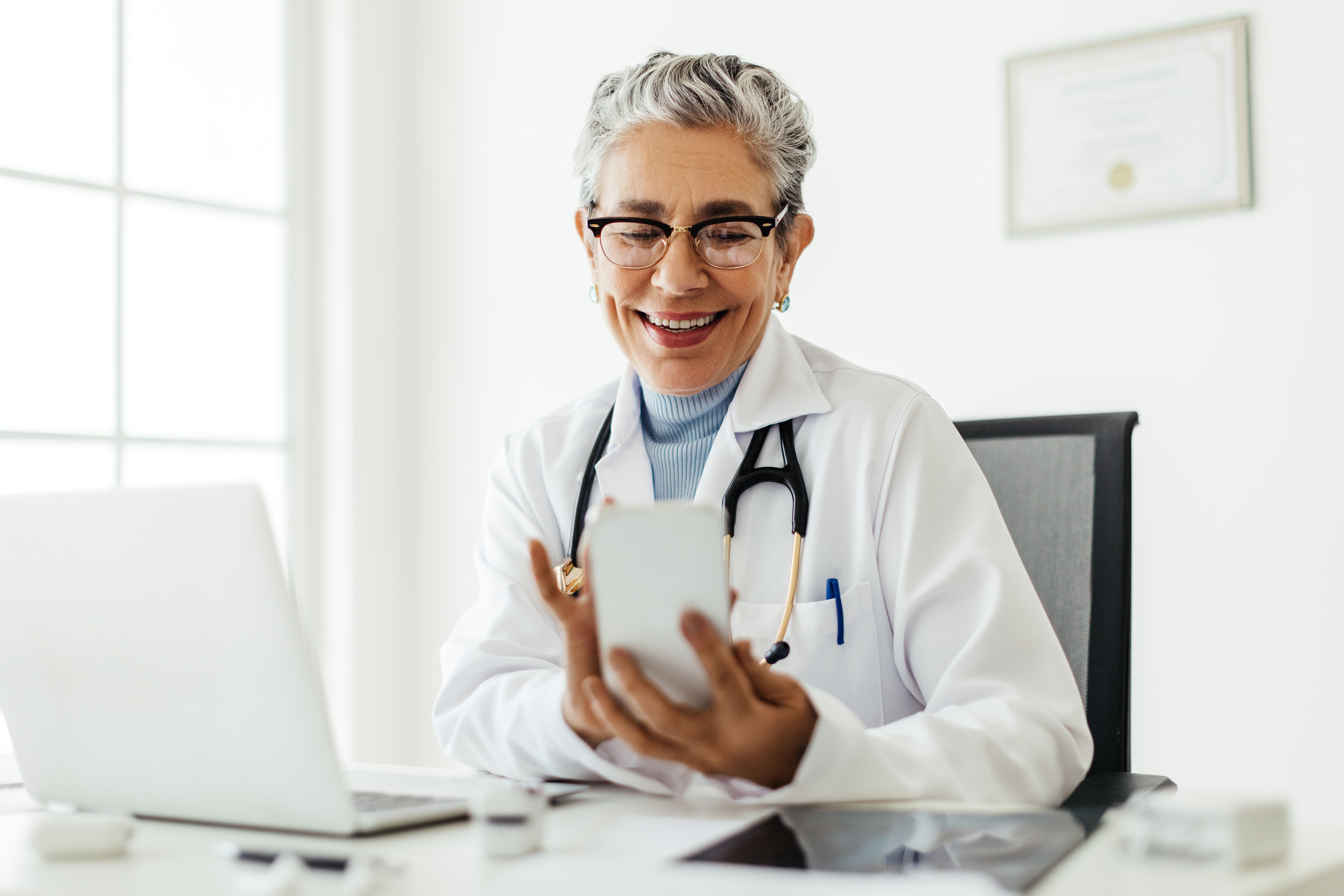female-european-elderly-doctor-consultation-room-using-her-cellphone-in-her-office-web